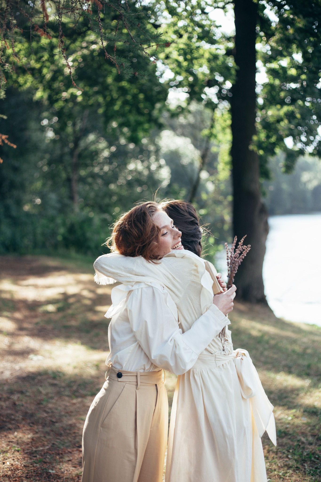 Two women in light dresses sharing an affectionate embrace beneath the trees; illustrative photograph